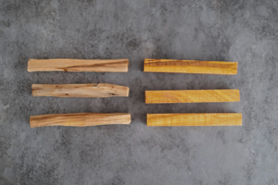 Three distilled palo santo sticks beside three resin-rich palo santo sticks, viewed from above on grey concrete